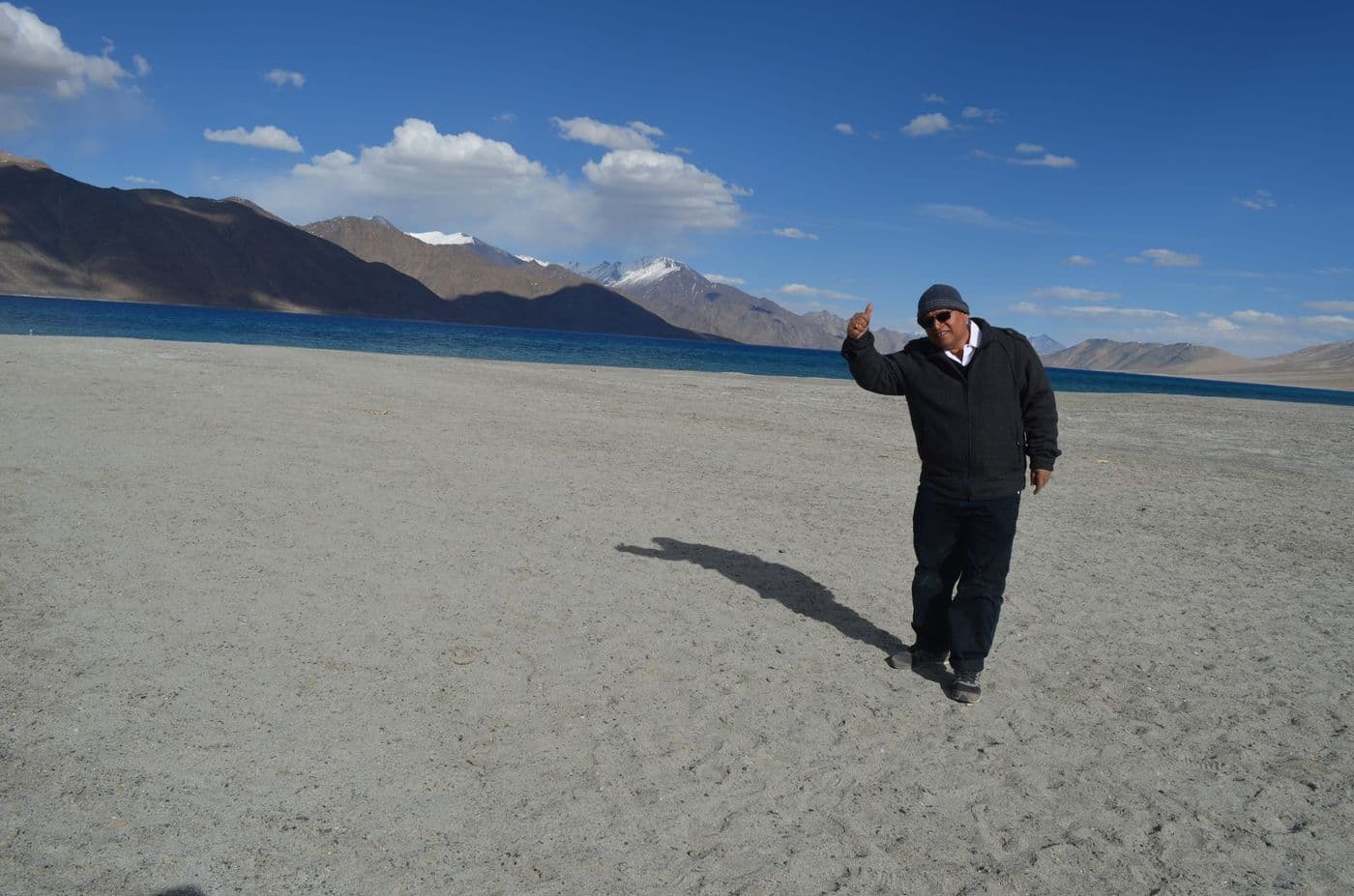 पैंगोंग झील के किनारे रेत पर खड़े होकर हाथ उठाए हुए, पीछे नीला पानी और बर्फ़ से ढके पहाड़